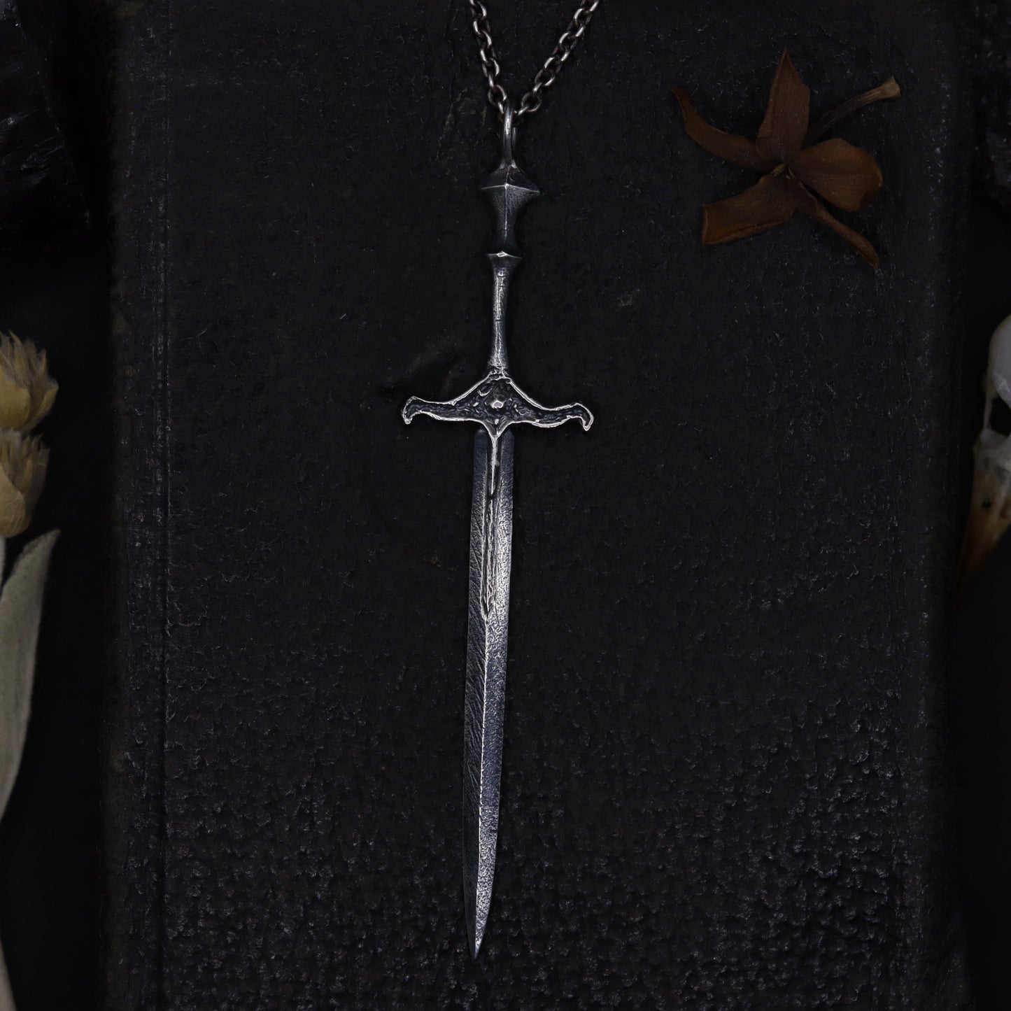 Silver sword-shaped pendant on a dark textured surface with dried plants and feathers.