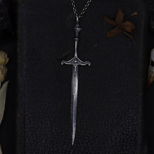 Silver sword-shaped pendant on a dark textured surface with dried plants and feathers.