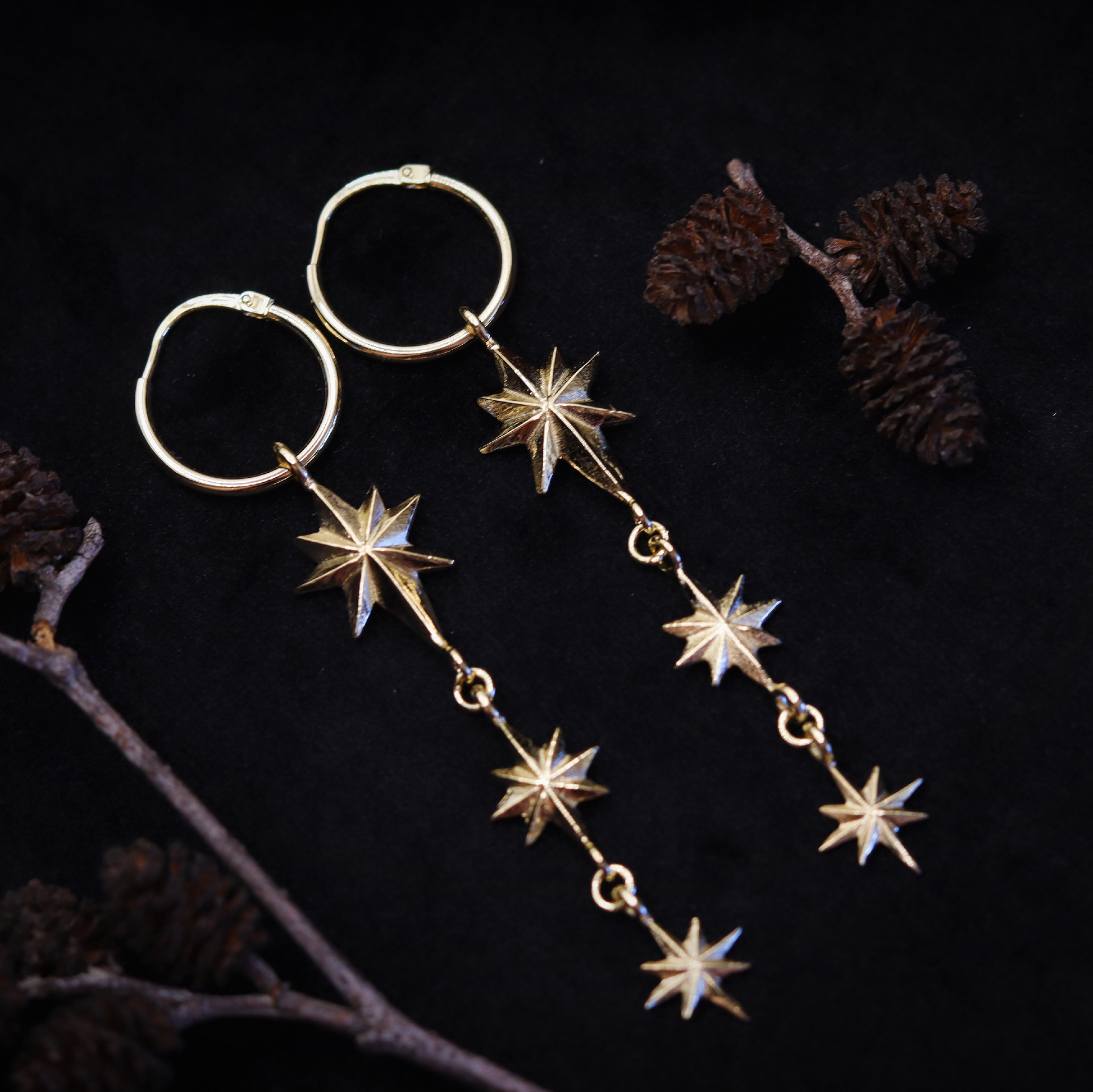 A pair of gold earrings featuring a series of star-shaped pendants on a black background, accompanied by pine cones and a branch.