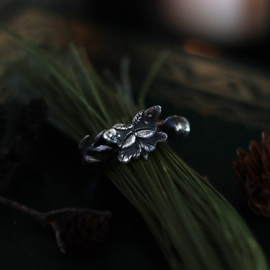A sterling silver ring with a butterfly and mushroom design, displayed on a dark background with pine needles and bits of mushroom.