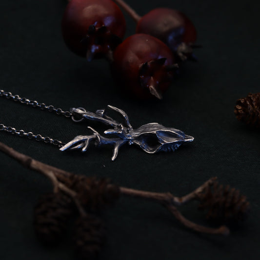A silver deer skull necklace on a dark background, surrounded by pine cones and berries.