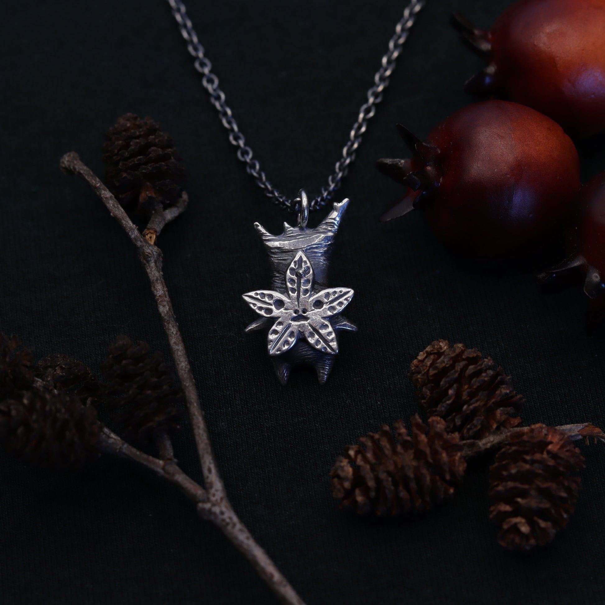 A silver necklace with a forest spirit pendant, displayed alongside pine cones and a branch on a dark background.