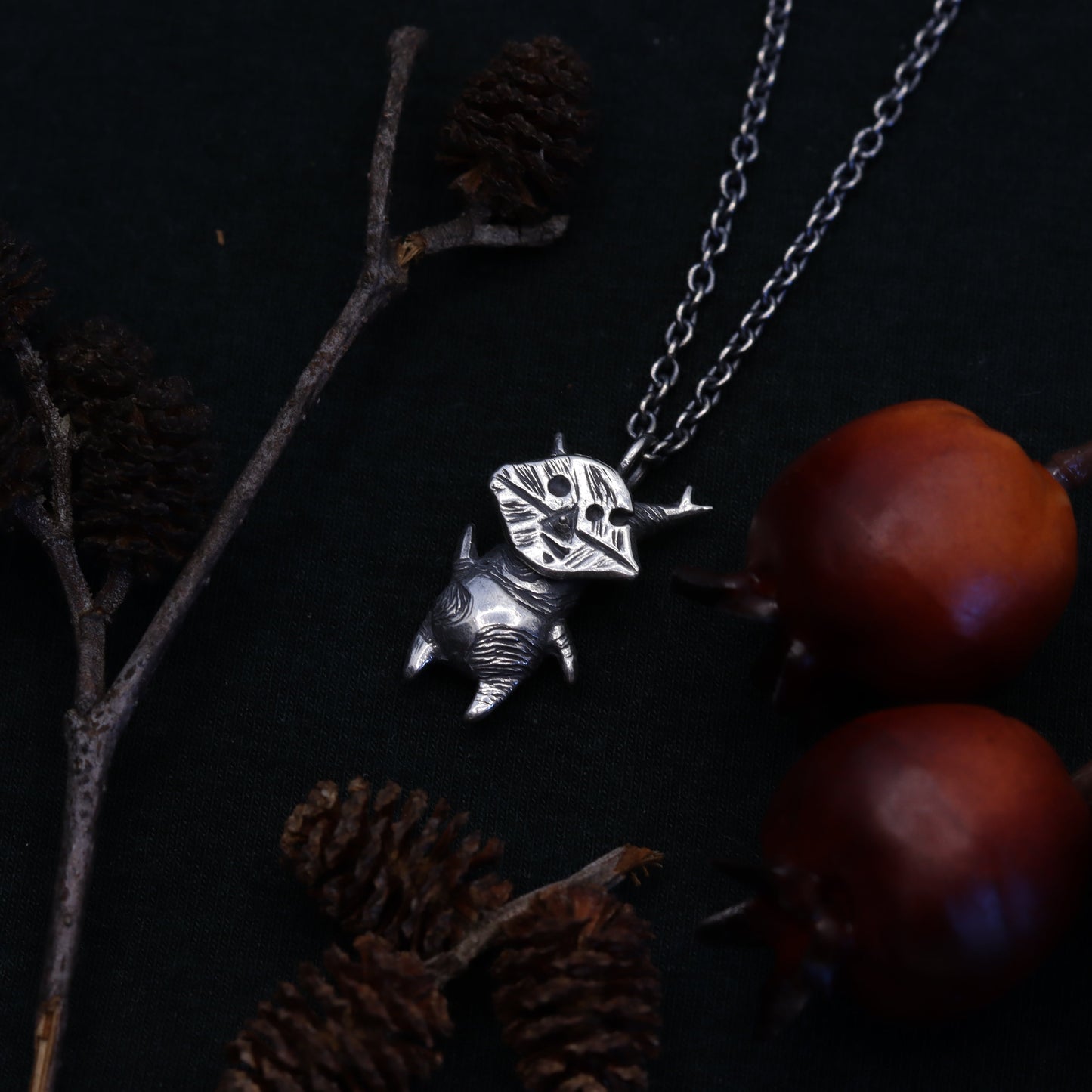 A sterling silver necklace with a forest spirit creature pendant, displayed against a backdrop of dark foliage and berries.