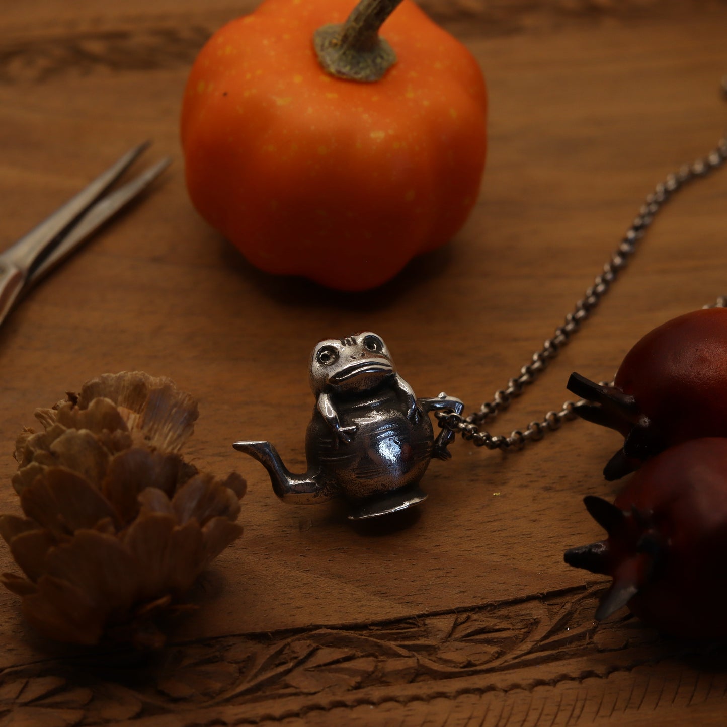 A necklace with a silver pendant shaped like a tea pot, displayed on a wooden surface with autumnal decorations including pine cones and a small pumpkin.
