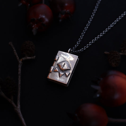 A silver necronomicon-inspired pendant necklace, displayed against a dark background with pine branches and red berries.