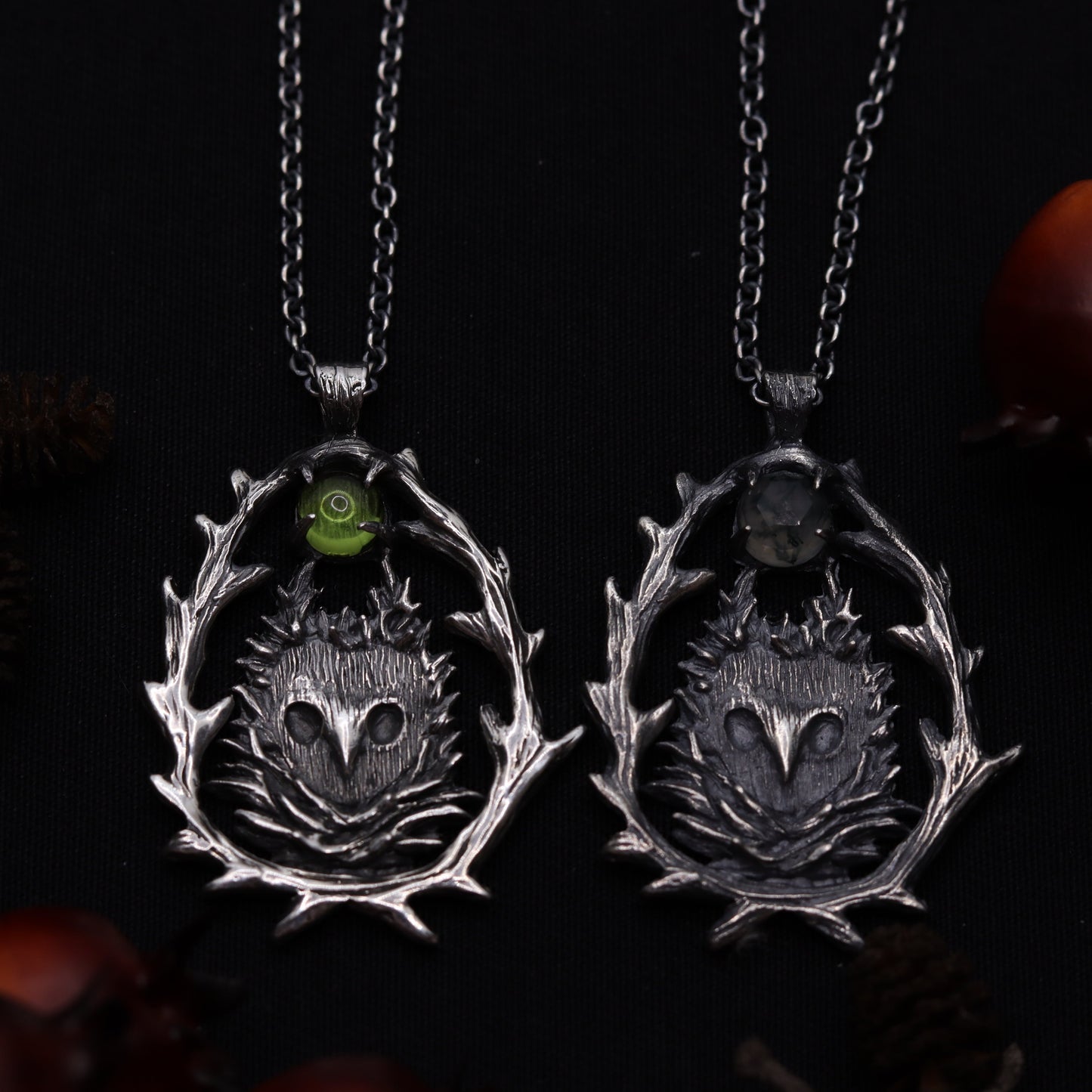 Two sterling silver owl-shaped pendants on chains, against a dark background with pine cones and decorative elements.