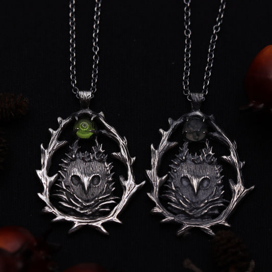Two sterling silver owl-shaped pendants on chains, against a dark background with pine cones and decorative elements.