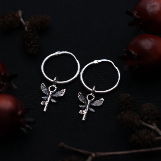 A pair of sterling silver earrings designed to resemble a key with wings, placed on a dark background with pine cones.