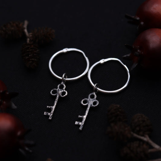 A pair of sterling silver earrings designed to resemble keys, displayed against a dark background with pine cones.
