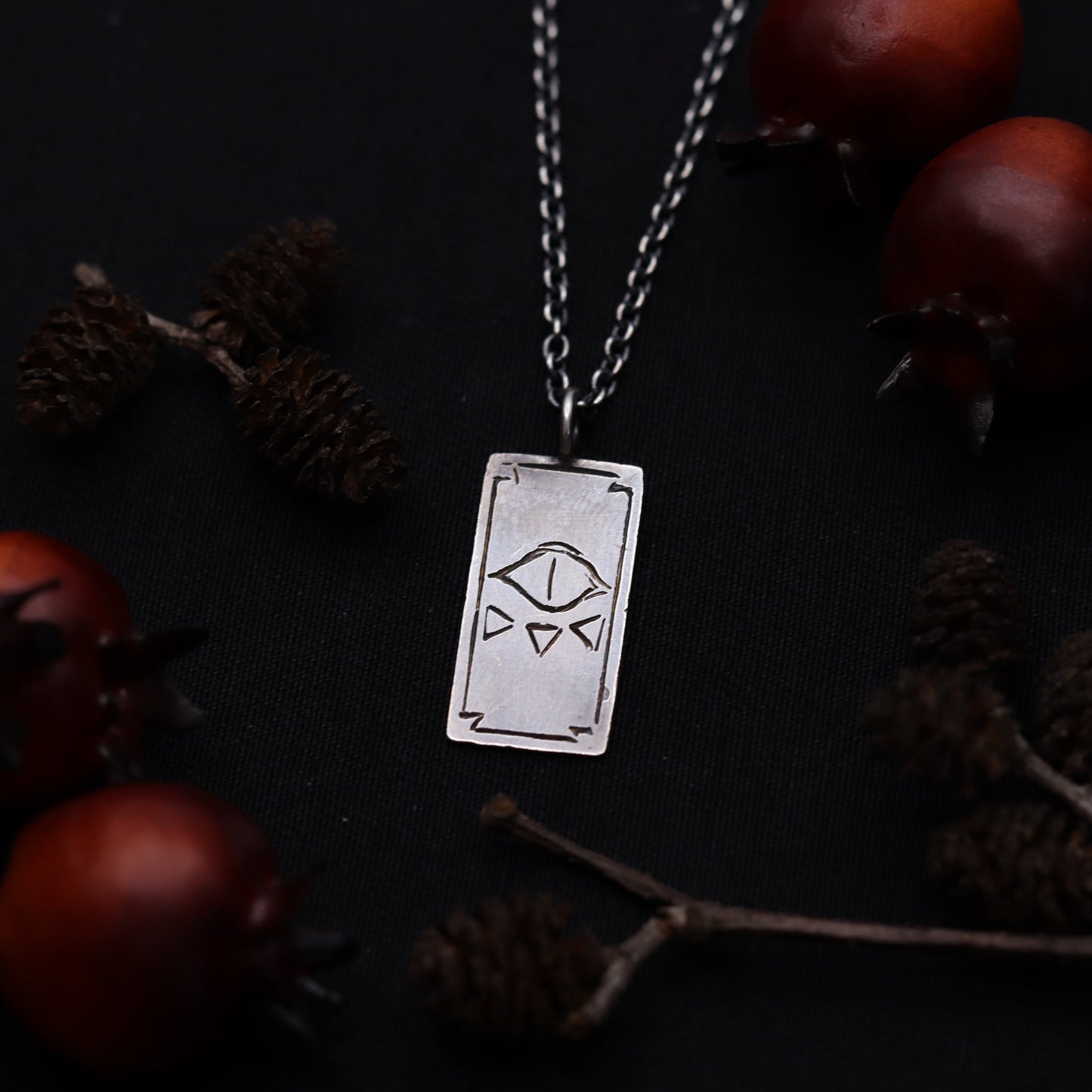 A silver necklace featuring a tarot card pendant with an evil eye design, displayed against a dark background with pine cones and red berries.