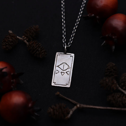 A silver necklace featuring a tarot card pendant with an evil eye design, displayed against a dark background with pine cones and red berries.