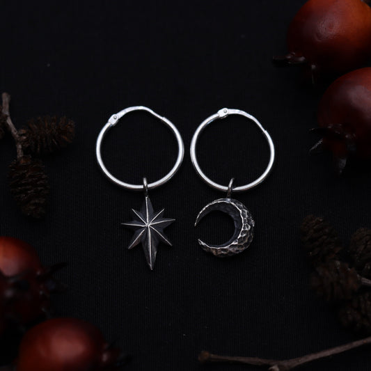 Delicate silver moon and star earrings laid on a dark surface surrounded by leaves and pine cones.