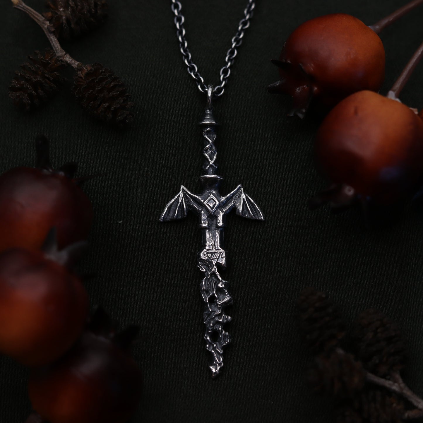A silvernecklace featuring a decorative sword design at its center, displayed on a dark background with pine cones around it.