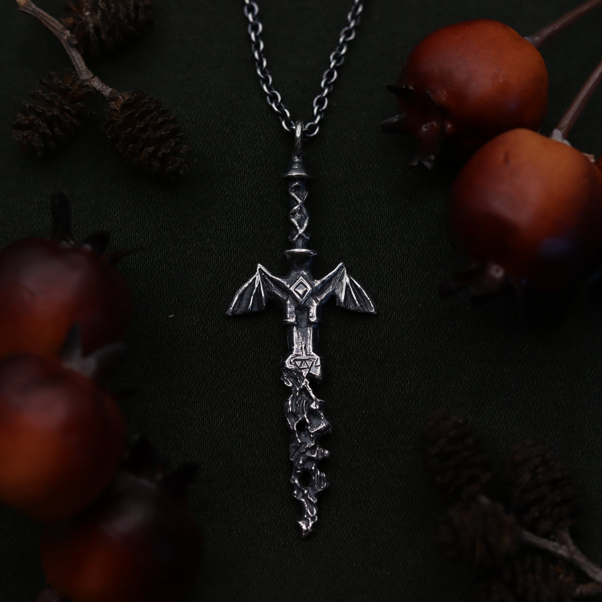 A silvernecklace featuring a decorative sword design at its center, displayed on a dark background with pine cones around it.