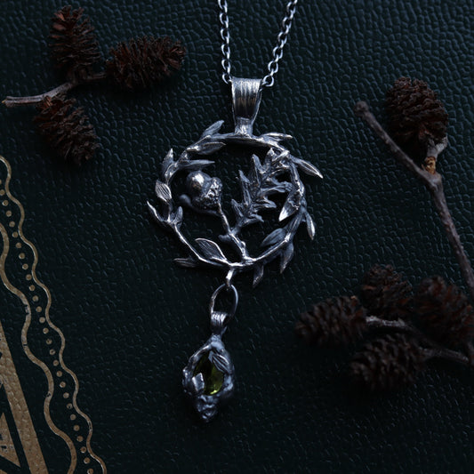 A sterling silver necklace with a peridot stone, designed as a talisman featuring an oak tree with branches and leaves, displayed against a dark background with intricate patterns.