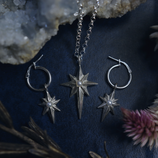A necklace and a pair of earrings featuring star-shaped designs, laid out on a dark textured surface with a crystal and some dried flowers nearby.