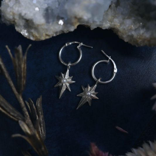 A pair of sterling silver earrings with star shapes and pearls at the center, displayed on a dark fabric with floral elements.