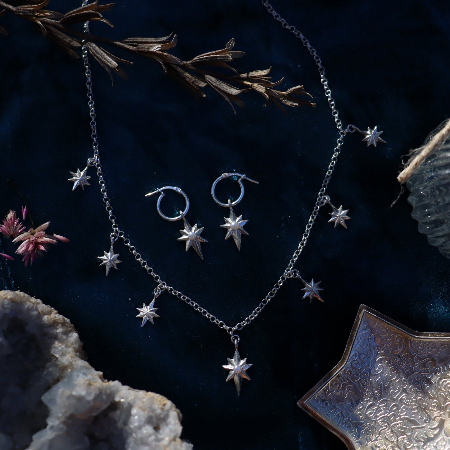 A jewelry set including a necklace and earrings with silver-colored stars and other geometric shaped charms, displayed on a dark background with a decorative rock and a sprig of dried flowers.