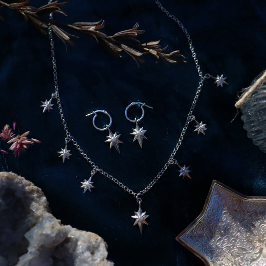 A jewelry set including a necklace and earrings with silver-colored stars and other geometric shaped charms, displayed on a dark background with a decorative rock and a sprig of dried flowers.