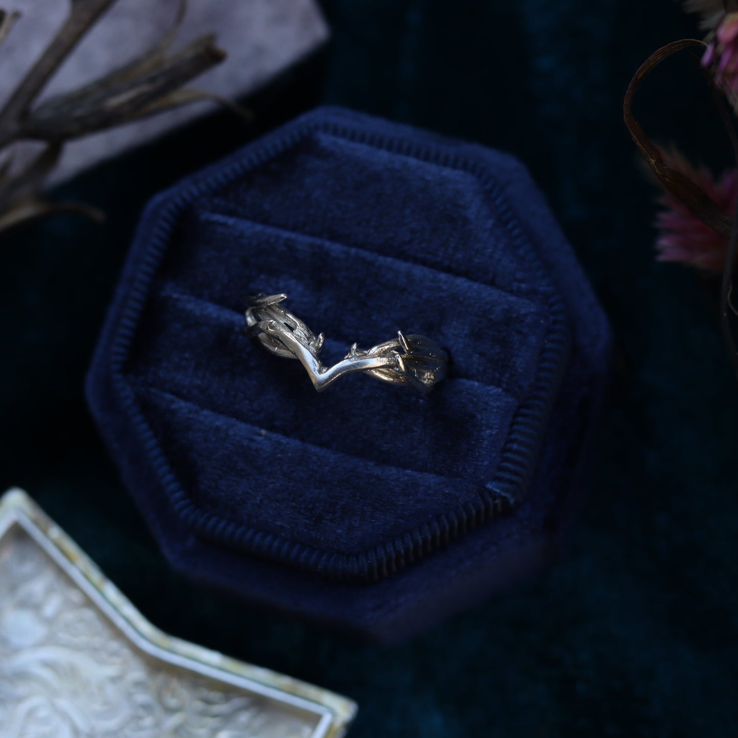 A sterling silver ring with a design resembling Galadriel's Circlet, displayed on a dark blue textured surface, surrounded by dried flowers and a small, square silver object.