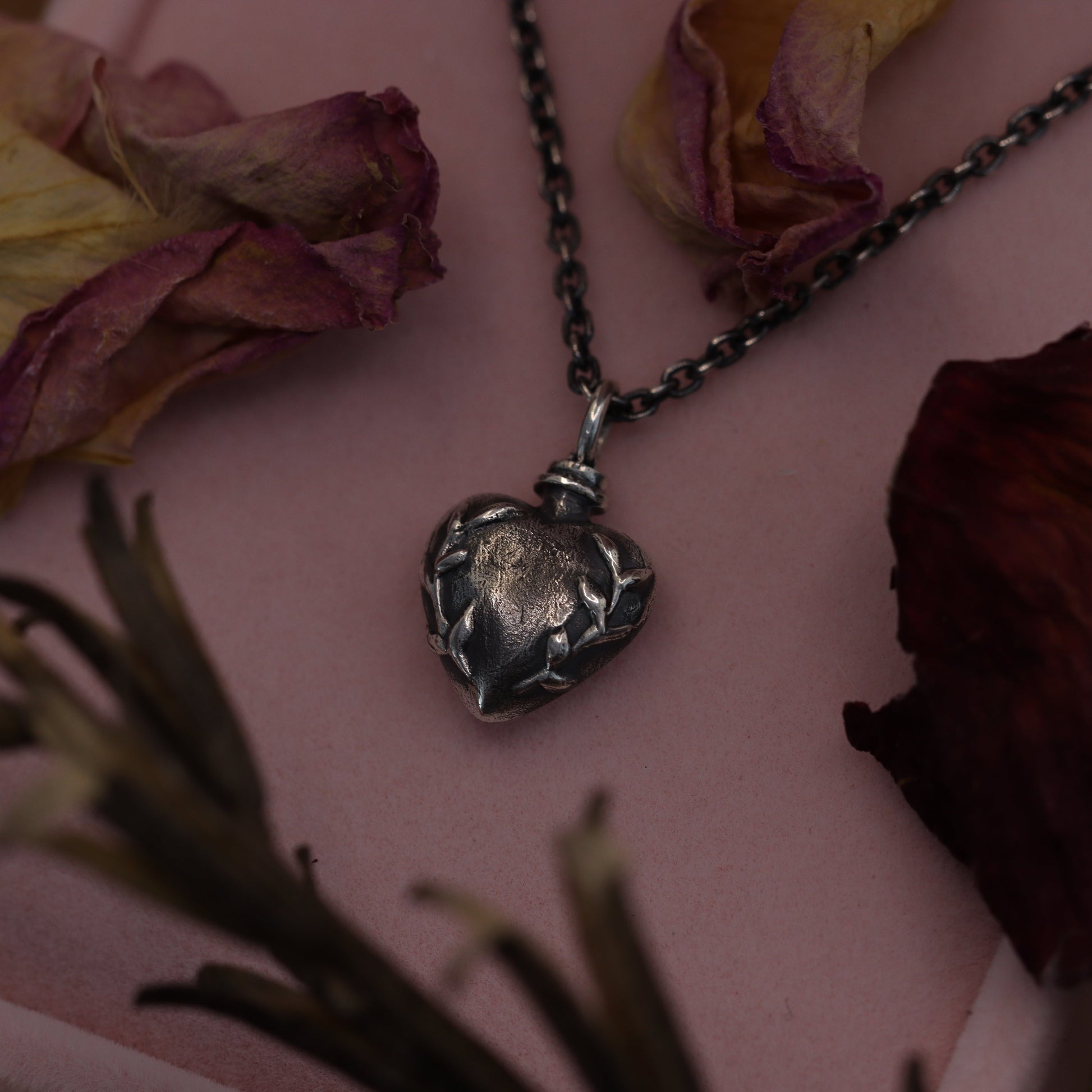 A sterling silver necklace with a heart-shaped love potion pendant, displayed on a pink surface with dried roses around it.