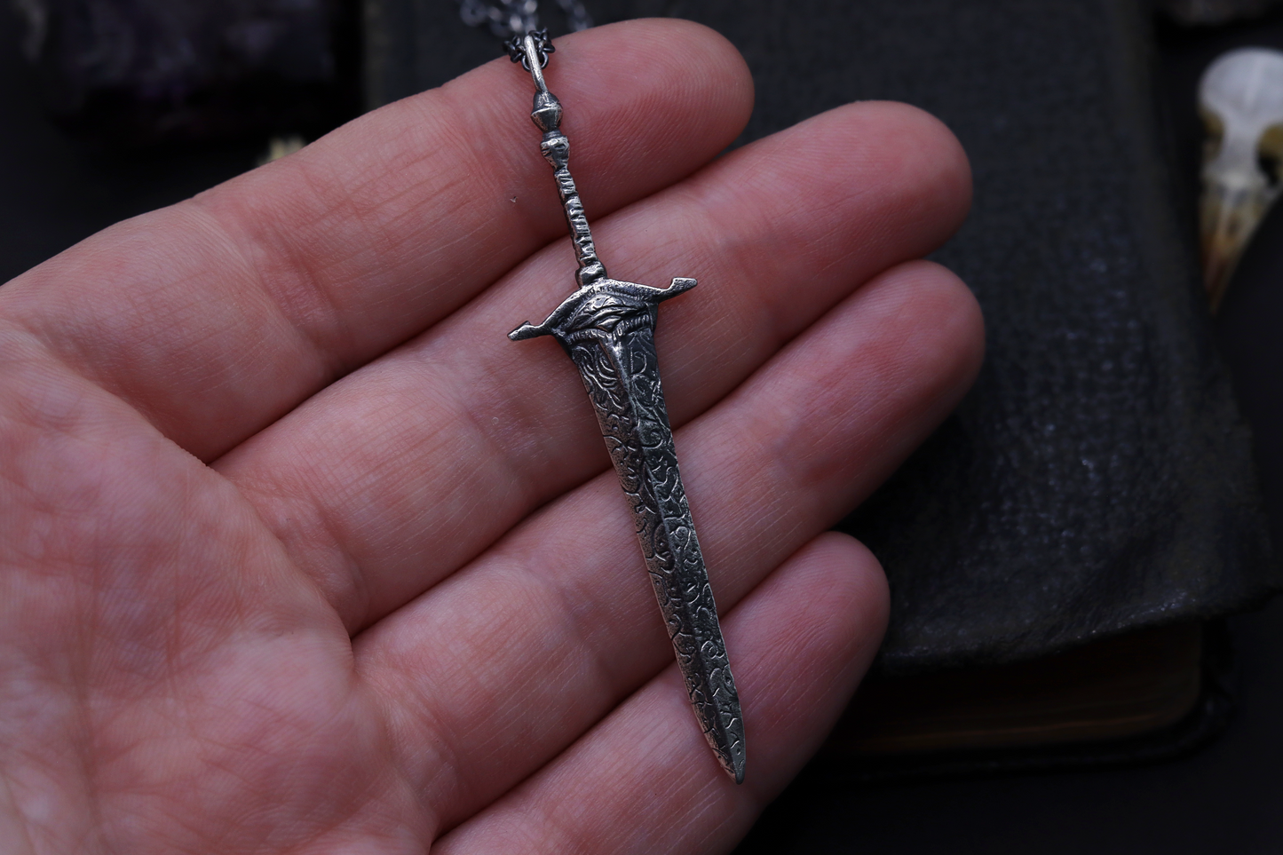 Silver sword-shaped pendant held in a hand against a dark background