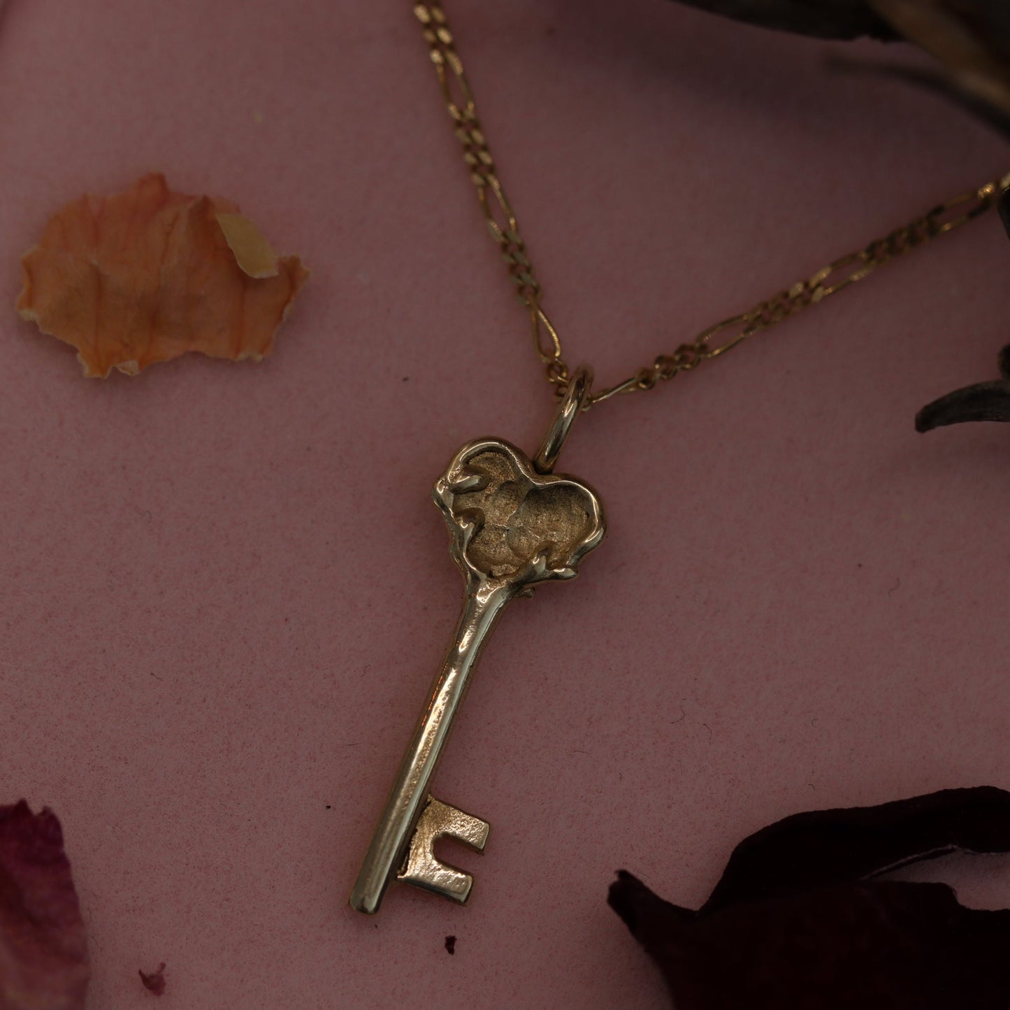 A 10k gold necklace with a key-shaped pendant, displayed on a pink background with dried flowers.