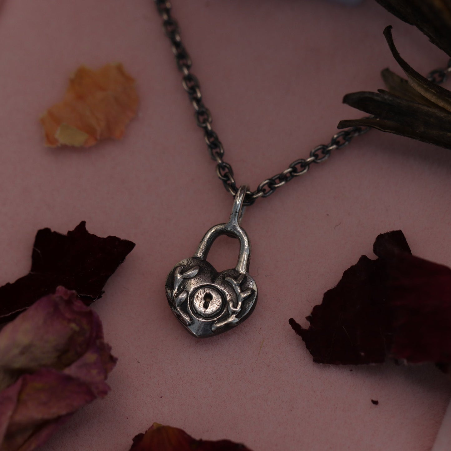 A sterling silver locket necklace with a heart-shaped design, displayed on a chain against a pink background with dried flowers.