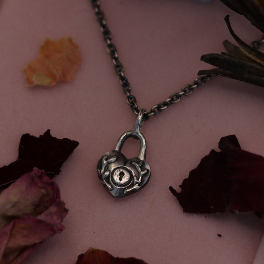 A sterling silver locket necklace with a heart-shaped design, displayed on a chain against a pink background with dried flowers.