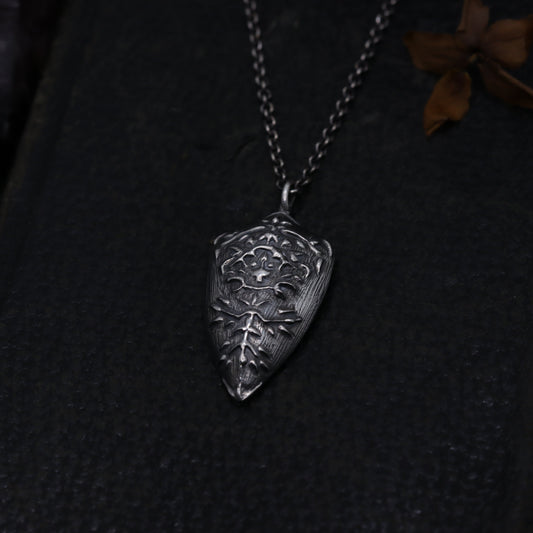 Silver Shield pendant necklace on a dark textured surface with dried flowers and a skull.