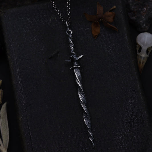 Silver sword-shaped pendant on a dark fabric background with dried plants.