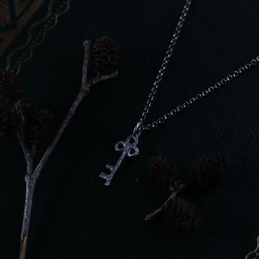 A sterling silver key-shaped necklace laid out on a dark surface with intricate patterns, with the chain partially visible.