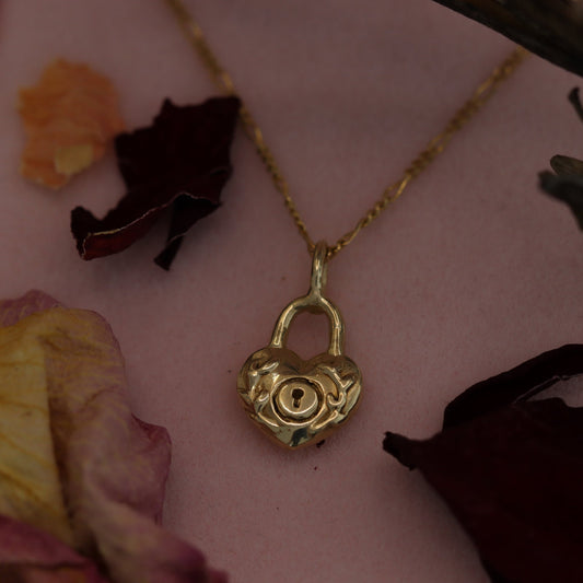 A 10K gold locket necklace displayed on a pink surface with dried flowers around it, the locket is engraved with a floral design.