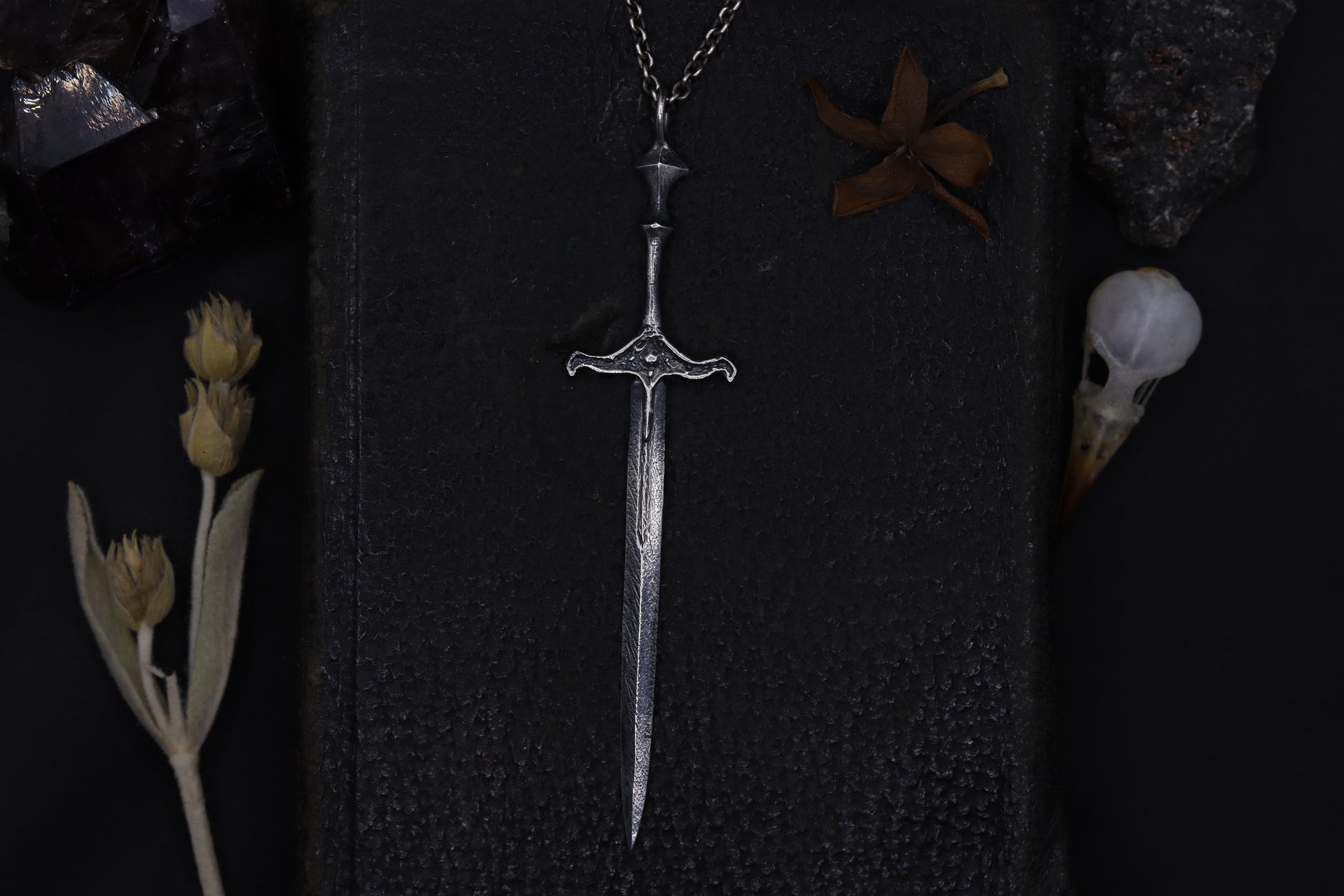 Silver sword-shaped pendant on a dark textured surface with dried plants and feathers.