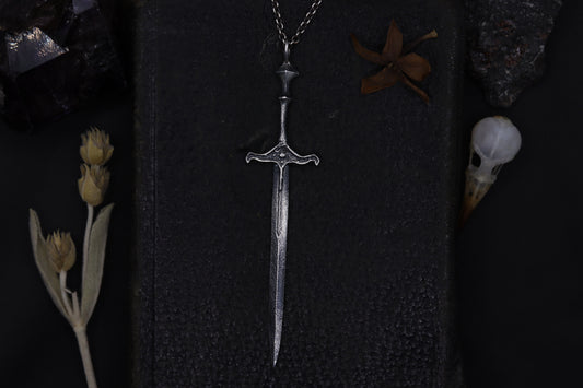Silver sword-shaped pendant on a dark textured surface with dried plants and feathers.