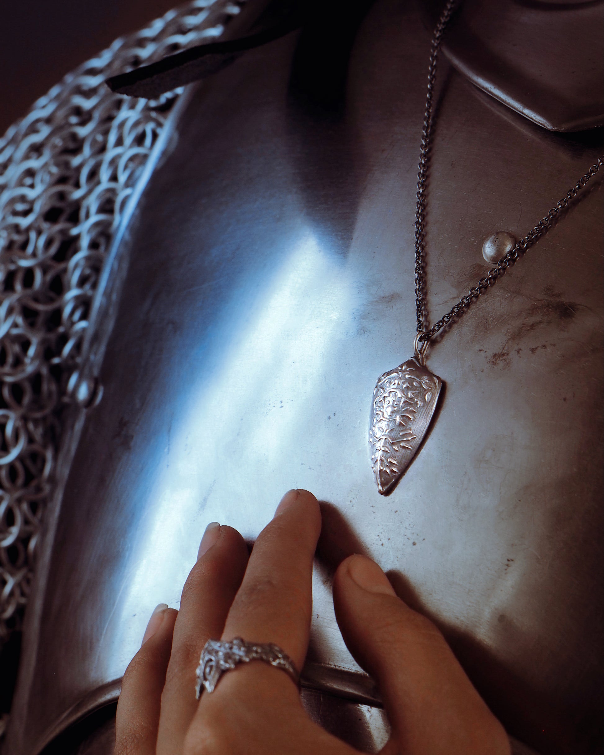 Silver necklace with a pendant on a metallic surface, with a hand wearing a silver ring.