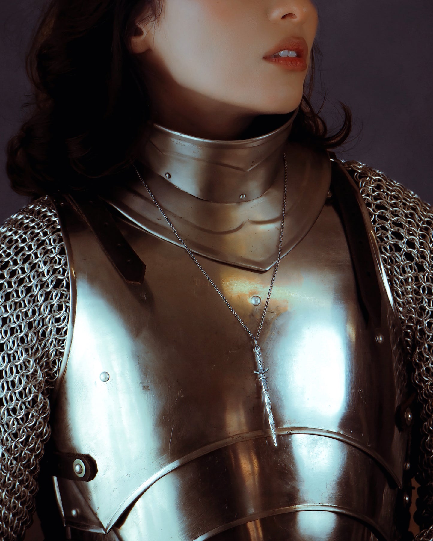 Person wearing chainmail armor with a sword-shaped necklace with a dark background
