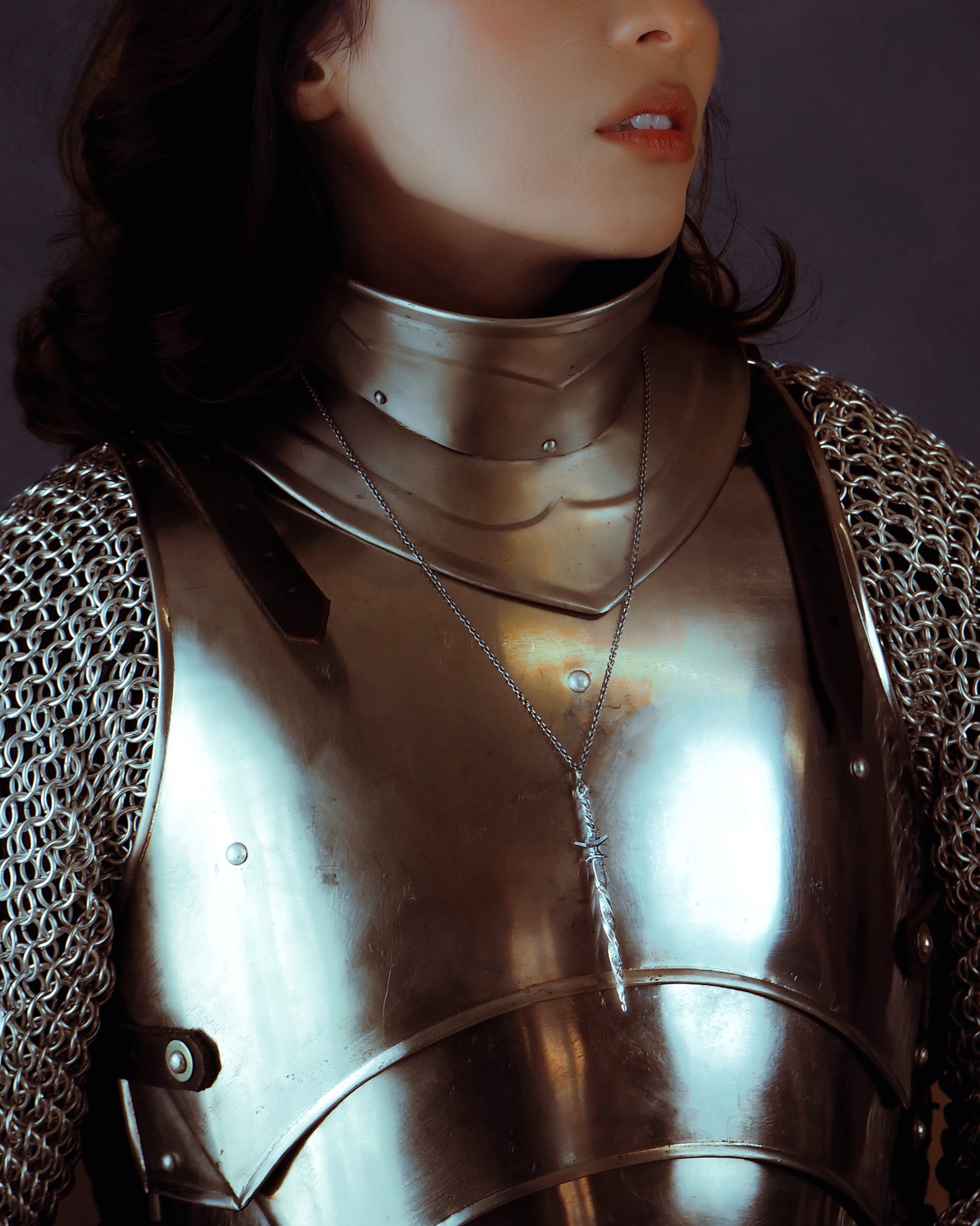 Person wearing chainmail armor with a sword-shaped necklace with a dark background