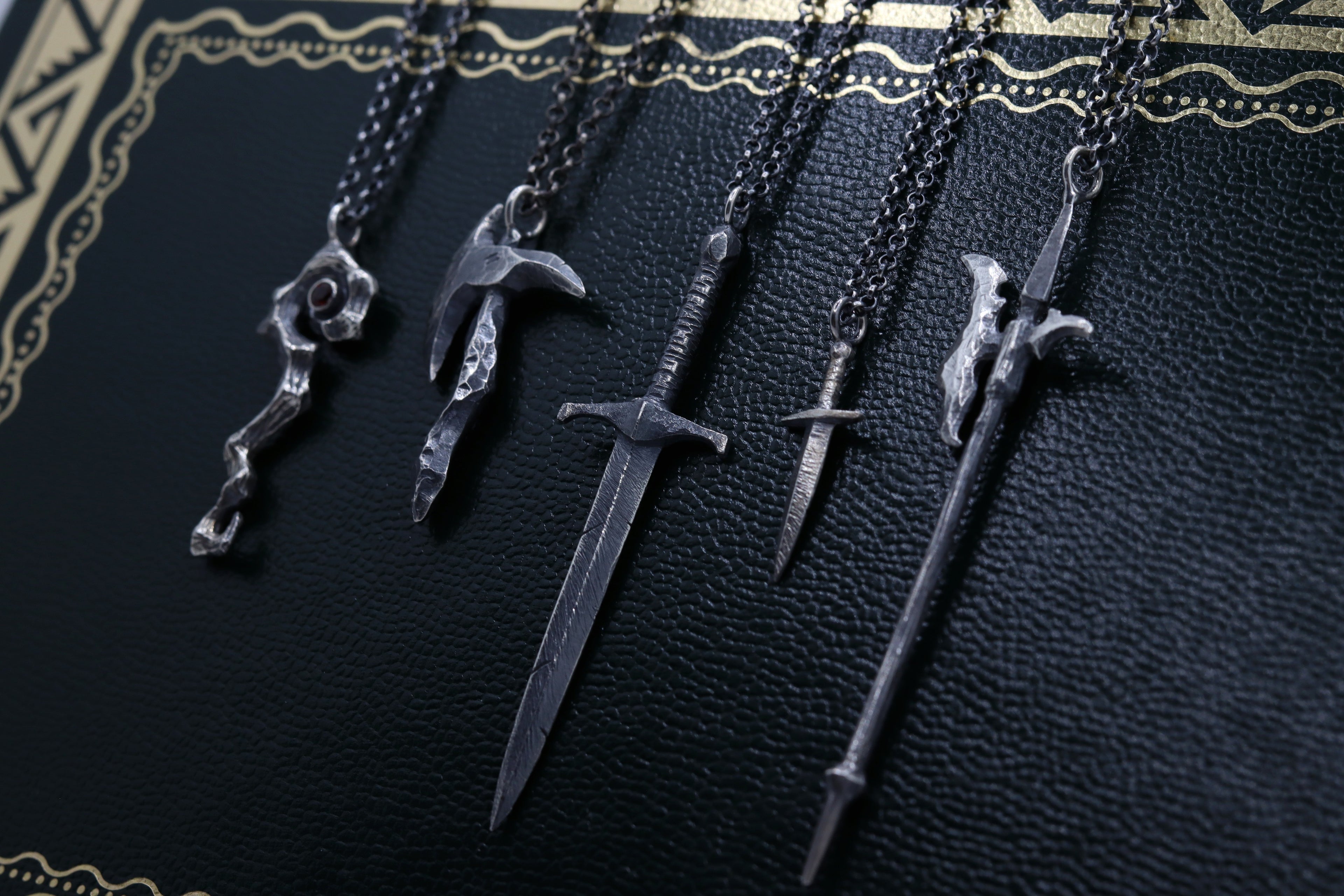 Hand-sculpted fantasy weapons, including a sword, staff, axe, dagger, and halberd, all crafted in sterling silver and hanging from a chain.