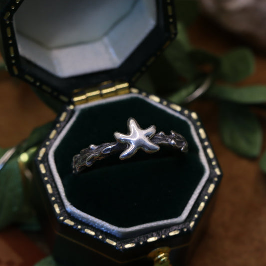 A ring with a star-shaped fruit design, placed inside an open black and gold box, with green leaves in the background.