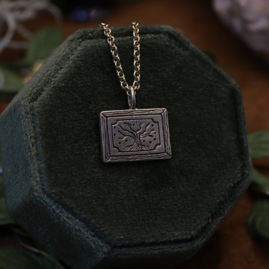 A necklace with a square pendant featuring a golden scroll design, displayed on a dark, textured surface with green leaves around it.