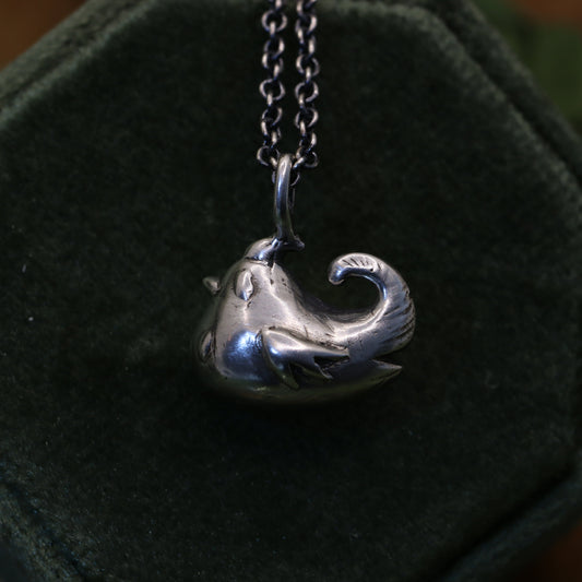 A pendant necklace featuring a hand-sculpted blue chicken, displayed on a black cushion.