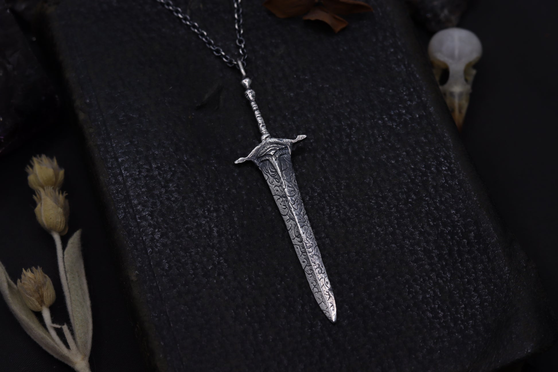 Silver sword-shaped pendant on a chain against a dark textured background with dried plants.