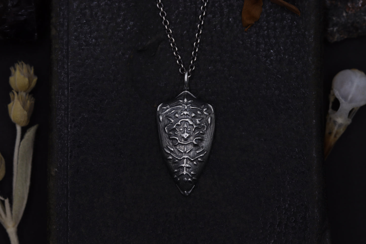Silver shield-shaped pendant on a dark textured background with dried flowers and a skull.