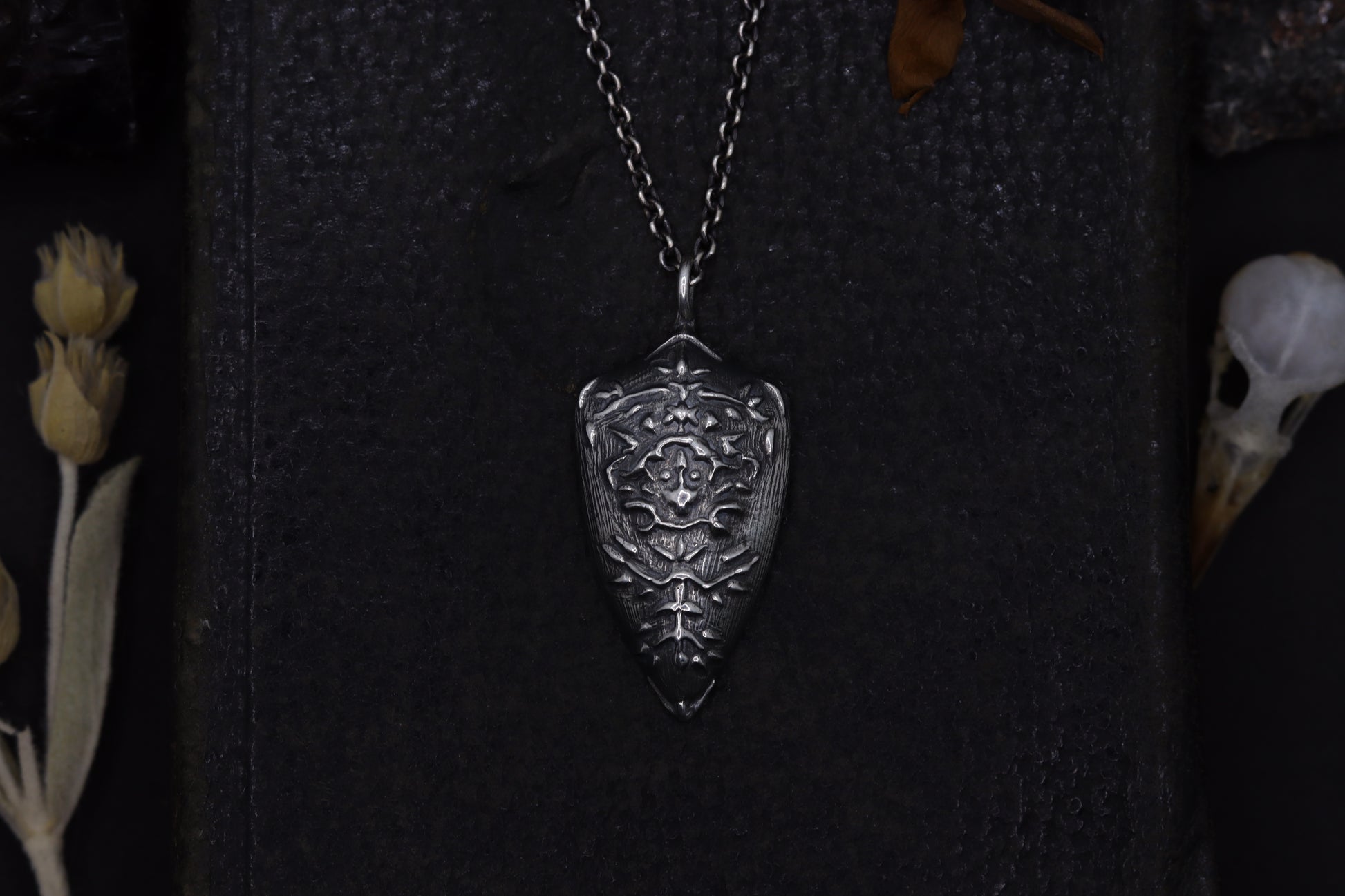 Silver shield-shaped pendant on a dark textured background with dried flowers and a skull.