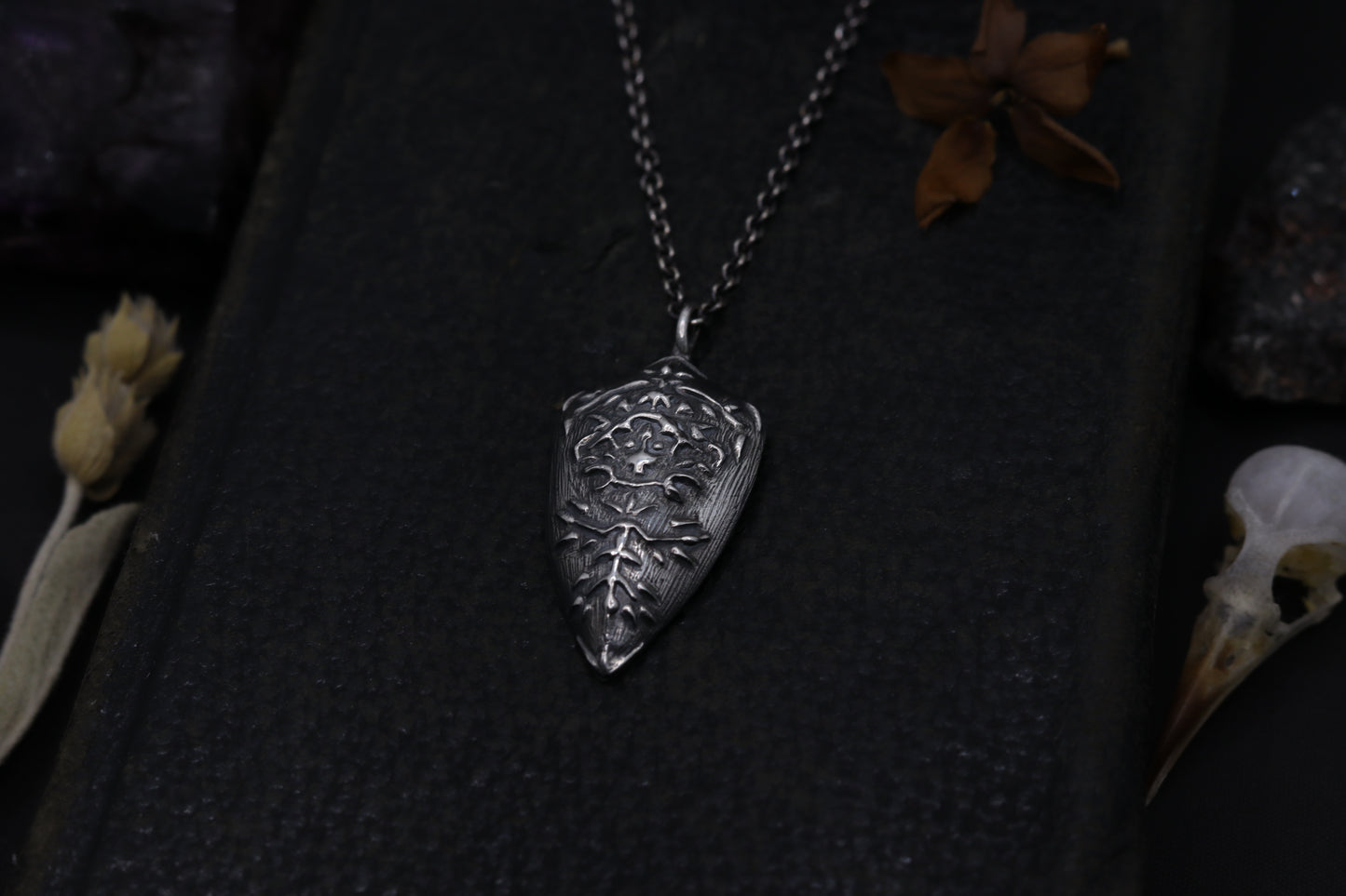 Silver Shield pendant necklace on a dark textured surface with dried flowers and a skull.