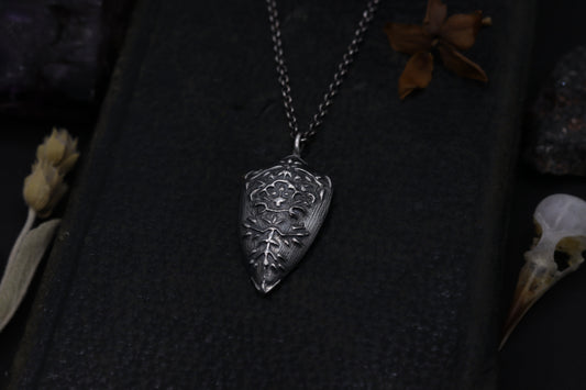 Silver Shield pendant necklace on a dark textured surface with dried flowers and a skull.