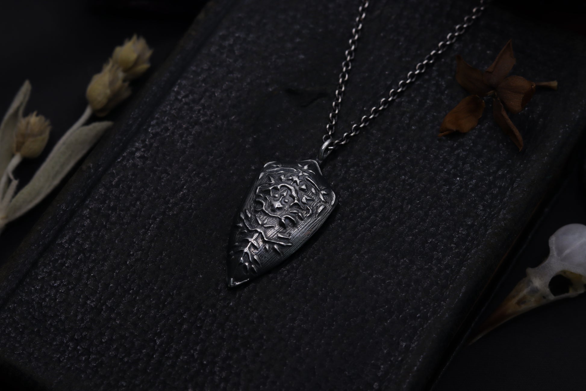 Silver necklace with a detailed shield-shaped pendant on a textured dark surface with dried flowers.