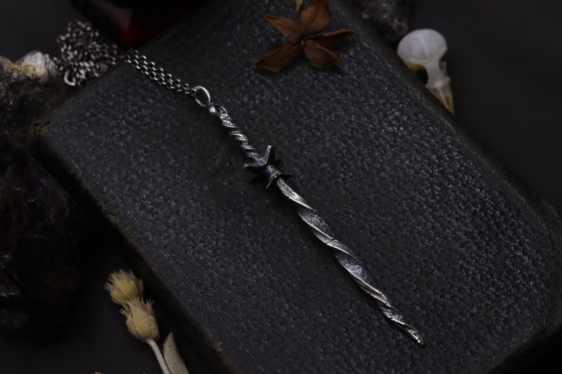 Silver sword-shaped pendant on a dark textured surface with dried flowers.